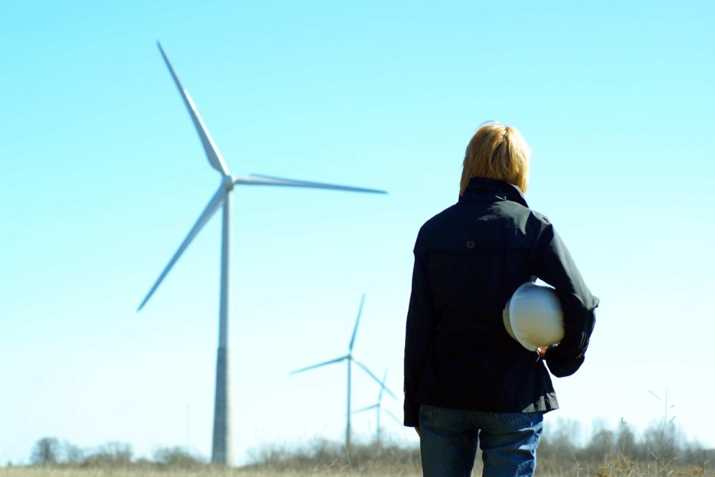 A woman carrying a hard hat inspecting a field with wind turbines