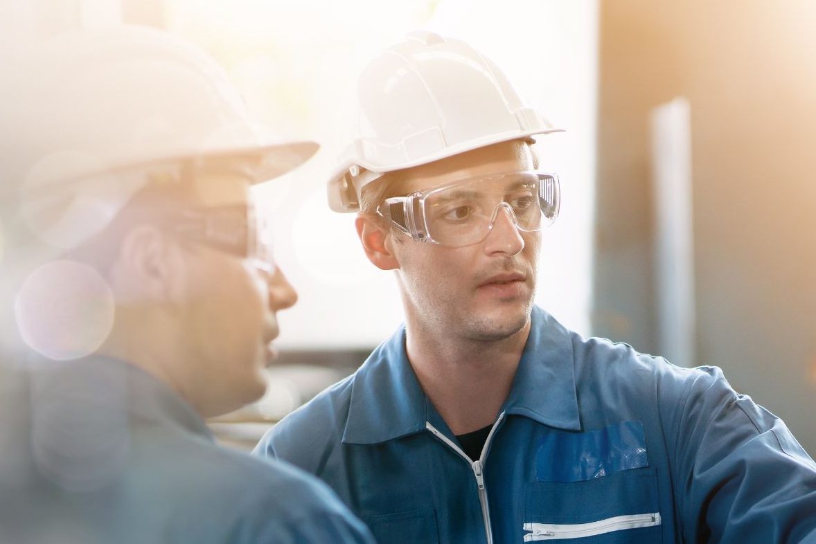 Two Individuals In Blue Overalls Talking To Each Other In An Industrial Setting Aspect Ratio 1200 800