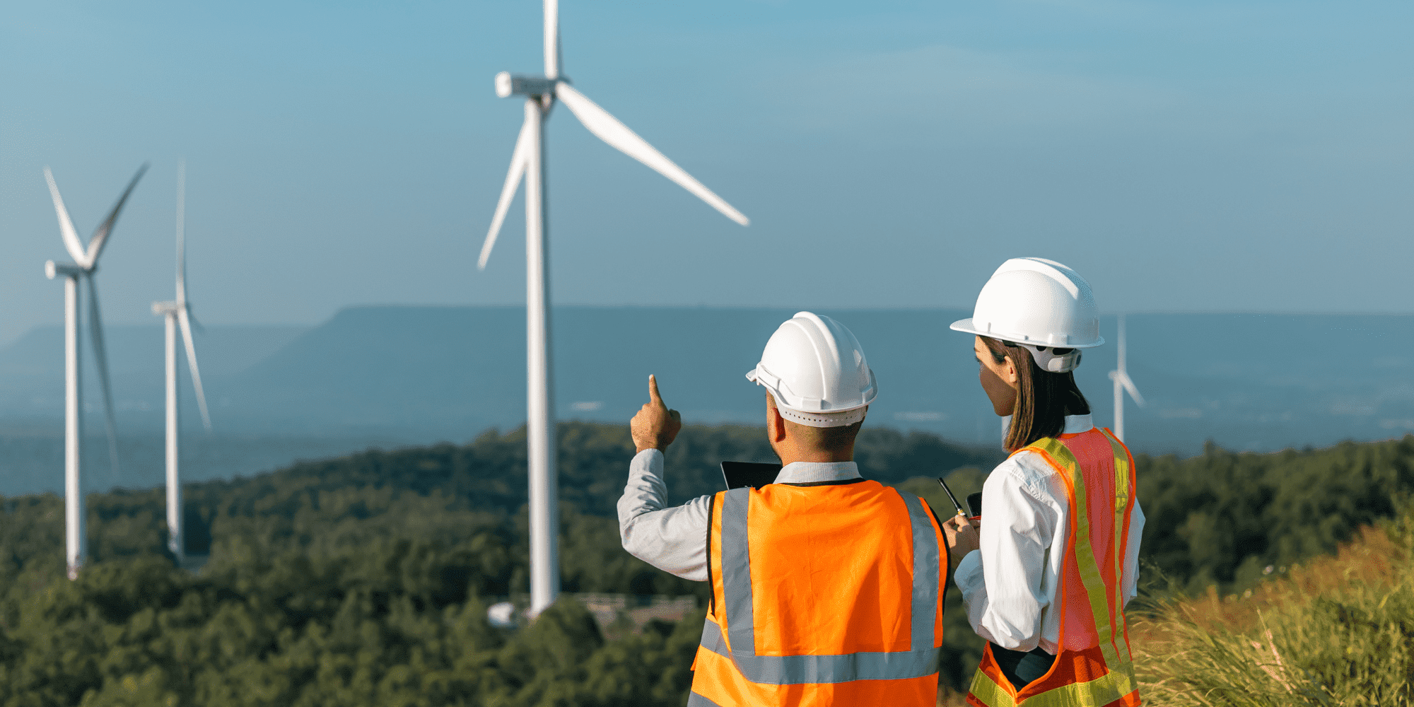 Wind Turbines A Lady And A Man In Orange Vests On A Hill Pointing At Wind Turbines Aspect Ratio 1160 580