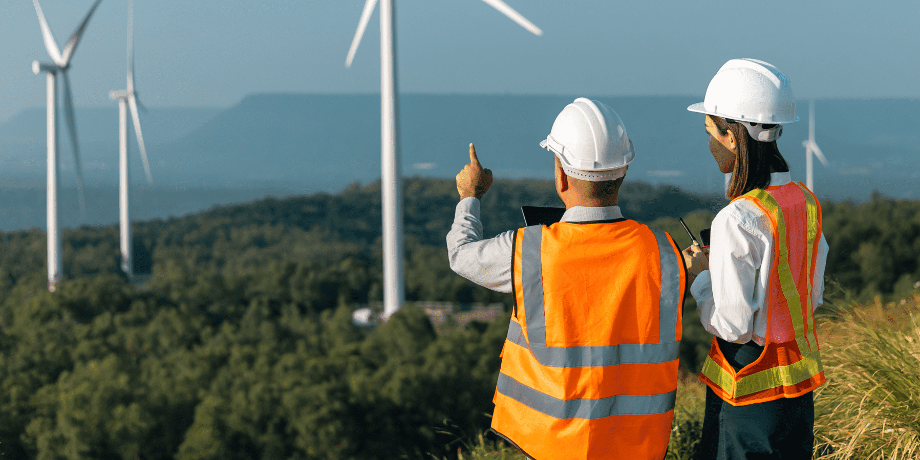 Wind Turbines A Lady And A Man In Orange Vests On A Hill Pointing At Wind Turbines Aspect Ratio 1160 580