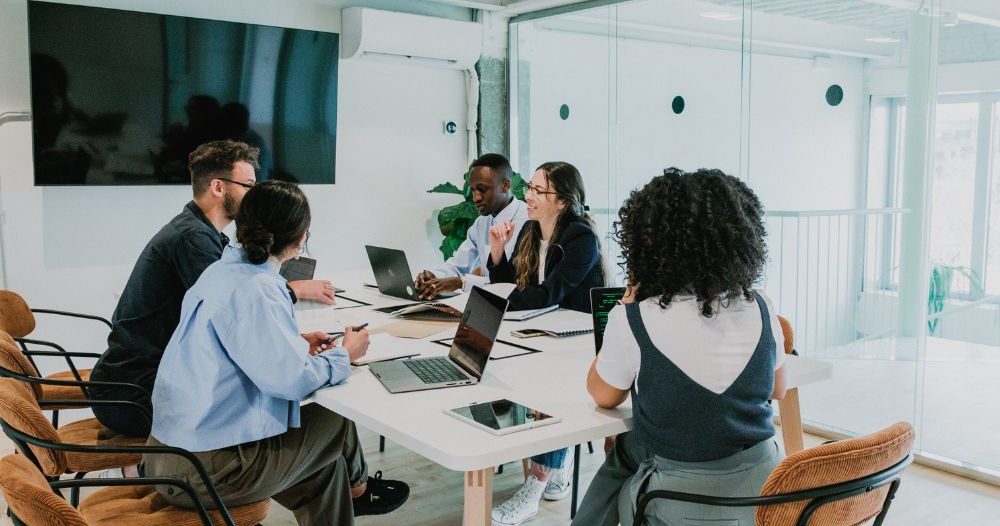 A Group Of People Sitting Around A Table In An Office Canva  Aspect Ratio 760 400